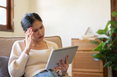 Düşünceli bir Asyalı kadın oturma odasındaki bir koltukta dijital tabletini kullanıyor. İnternetten ciddi videolar izliyor ya da internette çalışıyor..