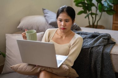 Güzel bir Asyalı kadın dizüstü bilgisayarında film izliyor ve yatak odasında dinlenirken kahvesini yudumluyor. İnsanlar ve eğlence kavramları