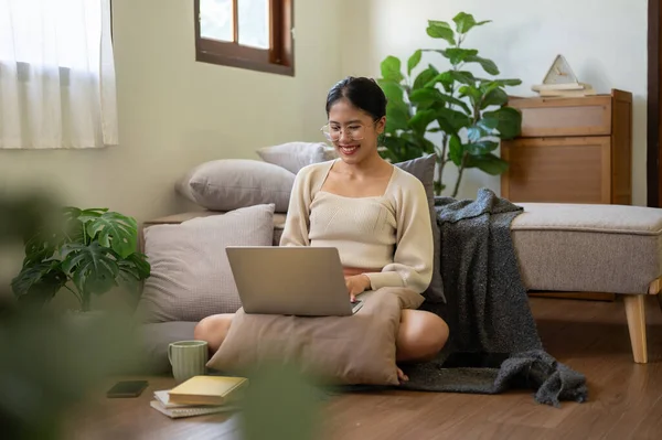 A beautiful and happy Asian woman is chatting with her friends on the internet via her laptop while sitting on the floor beside her bed in the bedroom. Domestic life, lifestyle, wireless technology