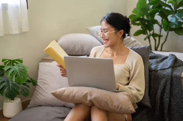 A happy and charming young Asian woman is working from home, researching information in a book, and working on her laptop while sitting in her bedroom.