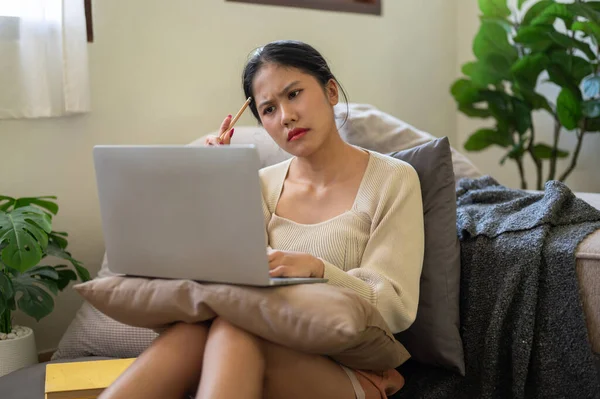 A focused and thoughtful Asian woman is working on her assignment on laptop in her bedroom, thinking and planning her work. work from home, domestic life