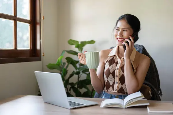 A beautiful Asian woman is sipping coffee while talking on the phone with someone at her desk in the home office. People and technology concepts