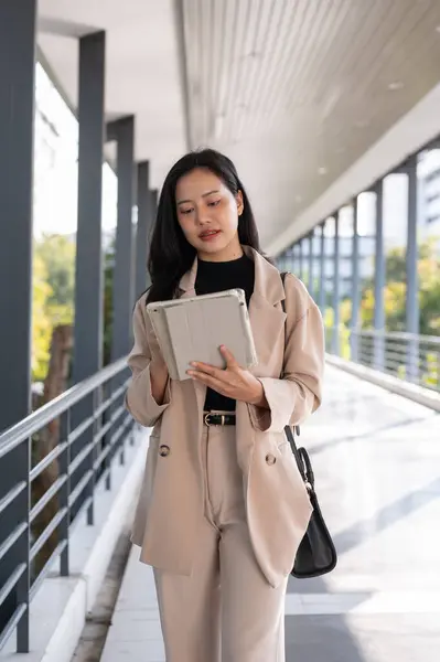 A professional and busy millennial Asian businesswoman focuses working on her work on her digital tablet while walking on a skywalk in the city.