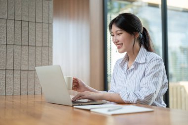 Çekici ve mutlu bir Asyalı iş kadınının yan görüntüsü bilgisayarındaki e-postalarına cevap veriyor ve modern bir iş yerinde ya da ofiste oturup kahvesini yudumluyor..