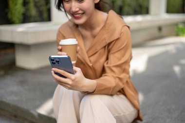 Çekici bir Asyalı kadının kırpılmış görüntüsü açık havada merdivenlerde oturuyor, akıllı telefonunu kullanıyor ve kahvesini yudumluyor, açık havada dinleniyor..