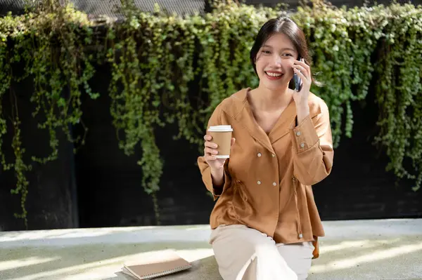 A beautiful and happy young Asian woman is enjoying talking on the phone with her friend and sipping coffee while relaxing on a bench in the garden on a bright day. People and lifestyle concepts