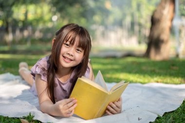 Güzel ve mutlu bir Asyalı genç kız, aydınlık bir günde yeşil parkta piknik battaniyesinde uzanırken kitap okumaktan zevk alıyor..