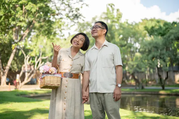 Lovely and happy Asian senior couples are holding hands and enjoying strolling around the green park on a bright day together. Happy marriage life concept