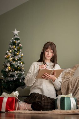 Rahat ve rahat elbiseli genç bir kadın Noel 'de oturma odasında dinlenirken günlüğünü yazıyor. Noel tatili ve ev hayatı kavramları