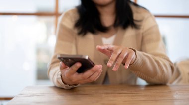 Kapalı bir masada otururken akıllı telefonunu kullanan bir kadın. Ekrana dokunmak, mobil uygulama kullanmak, mesajlaşmak, sosyal medyada kaydırmak. İnsanlar ve teknoloji kavramları