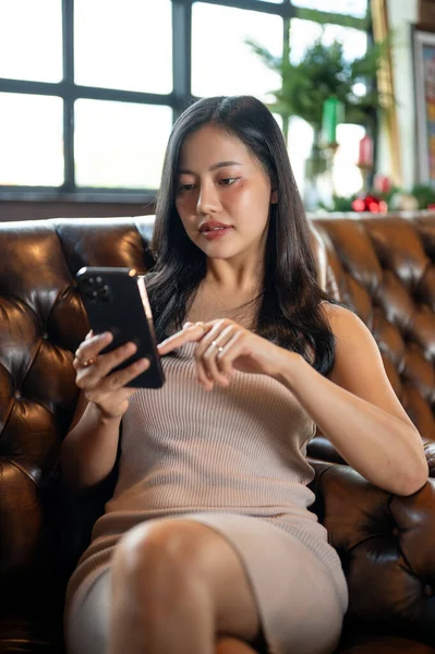 Young elegant Asian woman using smartphone in her free time while sitting in her expensive luxury sofa in her living room.