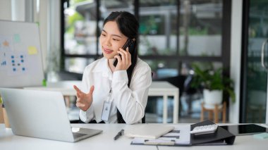 Asyalı iş kadını ofis odasında çalışırken müşterisiyle telefon görüşmesi yapıyor..