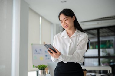 Modern ofis odasında dururken akıllı telefonu tutan ve kullanan iş kadını.