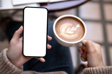 Bir kadının akıllı telefonunu kullanırken ve bir kafede açık bir masada otururken sabah kahvesinin tadını çıkarırken yakın plan görüntüsü. Beyaz ekran akıllı telefon modeli.