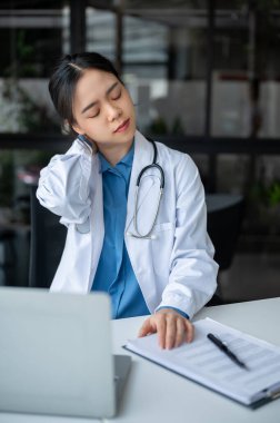 Yorgun ve çok çalışan genç bir Asyalı kadın doktor, hastane masasında çalışırken boynuna masaj yapıyor, boyun ağrısı ve ofis sendromu çekiyor..