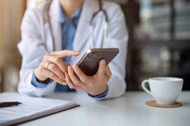 Hastane ofisindeki masasında akıllı telefonunu kullanan, mesajlara cevap veren ya da acil bir e-postayı kontrol eden bir kadın doktorun yakın plan görüntüsü..