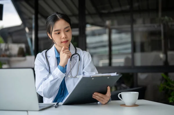 A professional and thoughtful young Asian female doctor is checking medical cases report on a clipboard and working in her office at the hospital.