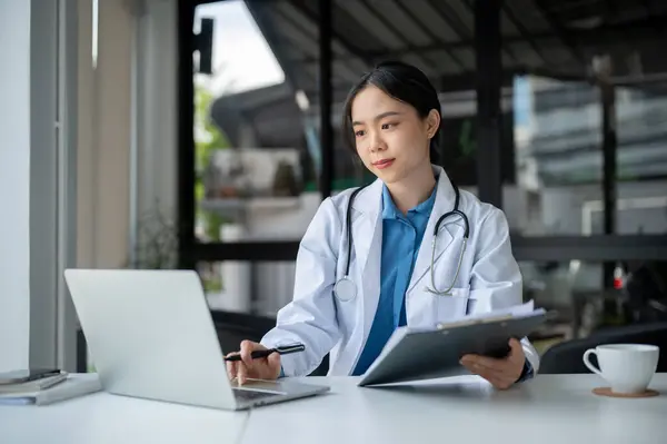 A beautiful and experienced young Asian female doctor in a white gown is working at her desk in the hospital, reading medical reports online.