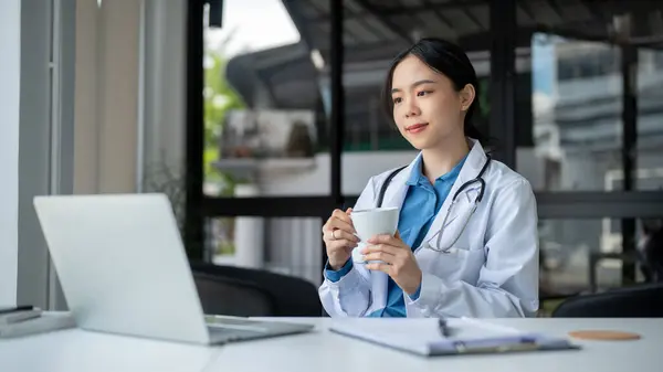 stock image A happy and successful young Asian female doctor in a white gown is enjoying coffee while working at her desk in the hospital. Health care and medical concepts