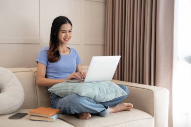 Genç ve güzel Asyalı kadın oturma odasındaki kanepede otururken dizüstü bilgisayar kullanıyor. Yaşam tarzı konsepti.