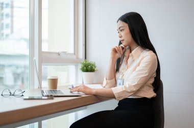 Odaklanmış ve profesyonel Asyalı bir iş kadını ofiste dizüstü bilgisayarı üzerinde çalışıyor. Yeni bir proje düşünüyor ve planlıyor..