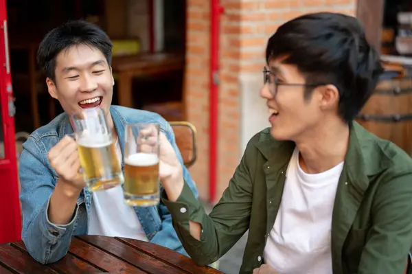 Two happy young Asian male friends are cheering and enjoying beers at a restaurant or bar in the city together. happy moment, hanging out, leisure, friendship