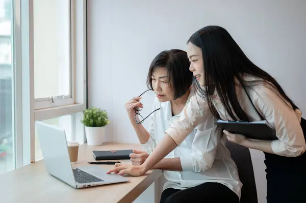 A beautiful young Asian businesswoman is helping a senior female colleague fix a computer\'s problem in the office.
