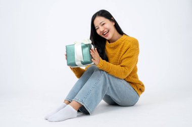 Hoş sarı süveterli, elinde sürpriz bir hediye kutusuyla beyaz bir stüdyonun arka planında, yerde oturan genç ve mutlu Asyalı bir kadın. Doğum günü hediyesi, Noel hediyesi.