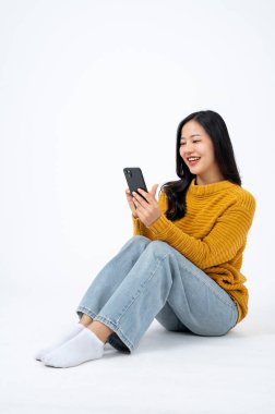 Genç ve güzel Asyalı kadın, günlük giysiler içinde yerde oturuyor ve akıllı telefonunu kullanıyor, izole edilmiş beyaz arka plan. sohbet, video çağrısı, sosyal medya