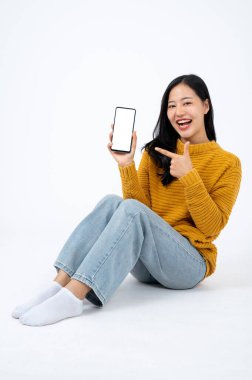 Gündelik giysiler içinde genç çekici bir Asyalı kadın izole edilmiş beyaz bir arka planda yerde oturuyor ve beyaz ekran akıllı telefon modeli gösteriyor..