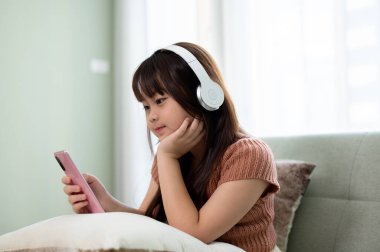 Günlük kıyafetlerle dolaşan, kulaklık takan ve oturma odasındaki bir kanepede akıllı telefonuyla çocuk çizgi filmlerini izleyen Asyalı genç bir kız. Çocuk ve teknoloji kavramları