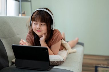 Kulaklık takan genç güzel Asyalı kız oturma odasındaki bir kanepede yatarken internetten çocuk videoları izliyor ya da dijital tabletinde ders çalışıyor. Teknoloji konsepti olan çocuk
