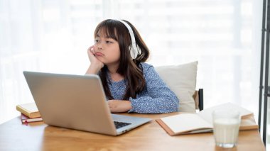 Sevimli genç bir Asyalı kız evde online çalışırken yorgun ve sıkılmış hissediyor. Çocuk ve online eğitim kavramları