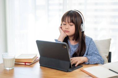 Kulaklık takan genç, sevimli bir Asyalı kız odasındaki bir masada dijital tabletinde ders çalışıyor. Çocuk ve online eğitim kavramları