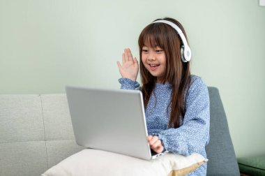 Kulaklık takan sevimli bir Asyalı kız oturma odasındaki kanepede otururken dizüstü bilgisayarda ders çalışıyor. Çocuk ve online eğitim kavramları