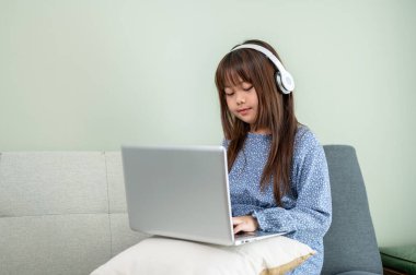 Kulaklık takan sevimli bir Asyalı kız oturma odasındaki kanepede otururken dizüstü bilgisayarda ders çalışıyor. Çocuk ve online eğitim kavramları