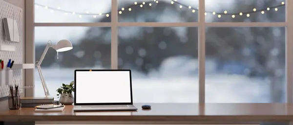 Home office workspace with a white-screen laptop computer and accessories on a wooden desk against the window with string lights on winter day. 3d render, 3d illustration