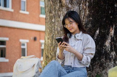 Olumlu ve çekici bir Asyalı bayan üniversite öğrencisi kampüs parkında ağacın altında dinlenirken akıllı telefonunu kullanıyor. sohbet, sosyal medyada kaydırma