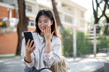 Şaşırmış ve heyecanlı genç bir Asyalı kadın telefon ekranına bakıyor, çarpıcı bir yüzle, ağzını kapatıyor, beklenmedik bir haber karşısında şaşırıyor, parkta bir bankta oturuyor..