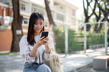 Sıradan giysiler içinde, pozitif ve çekici bir Asyalı kadın parkta bir bankta dinlenirken akıllı telefonuyla arkadaşlarıyla sohbet ediyor. İnsanlar ve teknoloji kavramları