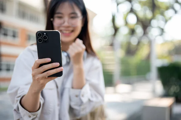 A cheerful young Asian woman is looking at her smartphone screen with a happy face and showing her fist while sitting on a bench in a park. good news, celebrating, lottery winning, selective focus