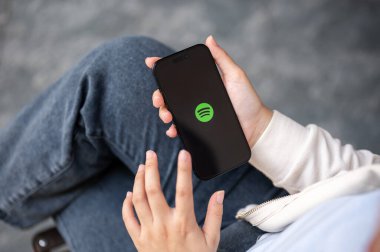 Chiang Mai, Tayland - 08 Aralık 2023: Açık havada dinlenirken akıllı telefonunda Spotify uygulaması kullanan bir kadın. Müzik uygulamaları, insanlar ve teknoloji kavramları