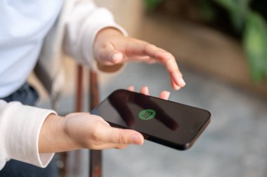 Chiang Mai, Tayland - 08 Aralık 2023: Açık havada dinlenirken akıllı telefonunda Spotify uygulaması kullanan bir kadın. Müzik uygulamaları, insanlar ve teknoloji kavramları