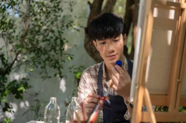 Önlüklü, pozitif ve yakışıklı Asyalı genç bir adam sanat eserini yaratıyor, bahçesindeki tuval sehpasında sulu boyayla resim yapıyor. Boş zaman ve yaratıcılık etkinliği kavramları