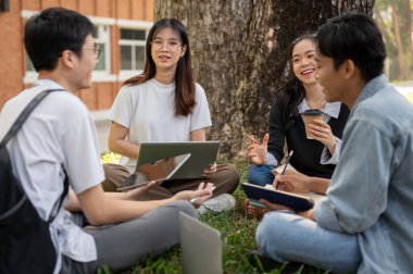 Bir grup mutlu genç Asyalı öğrenci kampüs parkında çimlerin üzerinde otururken ev ödevi yapıyor ve konuşuyor. Eğitim ve arkadaşlık kavramları