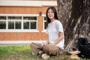 Gözlüklü, pozitif ve gülümseyen Asyalı genç bir üniversite öğrencisi dijital tabletiyle ağacın altında oturuyor ve kampüs parkında dinleniyor..