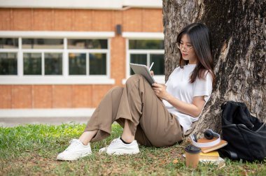 Güzel ve rahat bir Asyalı kız üniversite öğrencisi kampüs parkındaki ağacın altında dijital tabletini kullanıyor. İnsanlar ve teknoloji kavramları