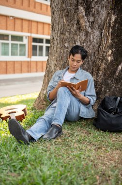 Sıradan kıyafetli genç bir Asyalı erkek üniversite öğrencisi kitap okuyor ya da kampüs parkındaki ağacın altındaki bir kitaba bazı fikirler yazıyor. Üniversite hayatı, yaşam tarzı, eğlence.