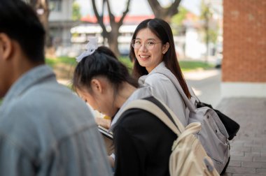 Olumlu, çekici Asyalı genç bir üniversite öğrencisi kameraya gülümsüyor, kampüste arkadaşlarıyla birlikte bir patikada oturuyor. Okula, üniversite hayatına, okul kızına, arkadaşlığa