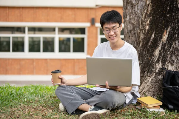 年轻的亚洲男大学生正在校园公园的一棵树下使用他的笔记本电脑 大学生活 人和无线技术概念 — 图库照片